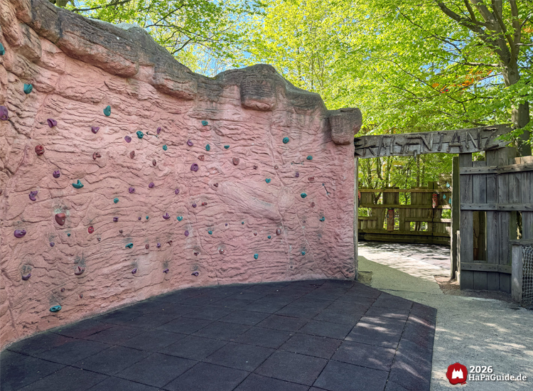 Kletterwand im Abenteuerland des Hansa-Park mit roter Felsstruktur, bunten Griffen und Eingang zum Regentanz unter grünen Bäumen