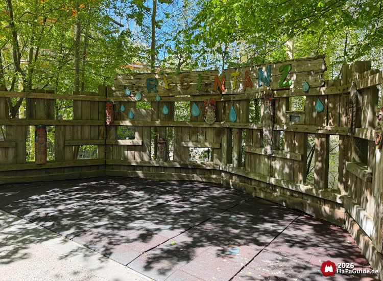 Holzmauer im Abenteuerland des Hansa-Park mit buntem Regentanz-Schriftzug, bemalten Masken und blauen Regentropfen Sprungmatten unter schattigen Bäumen