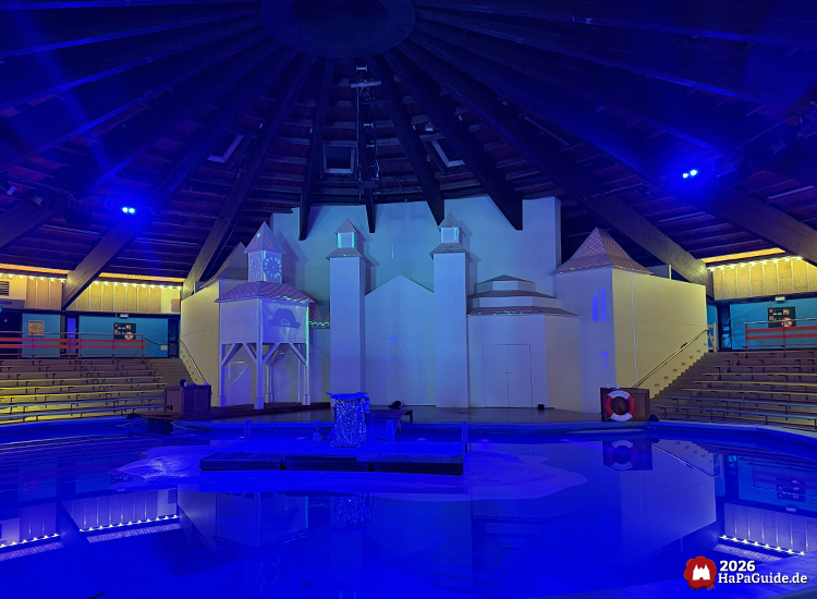 Innenraum der Aquarena im Hansa-Park mit blauer Beleuchtung, Wasserbecken und weißer Bühnenkulisse mit Türmen und Pavillon