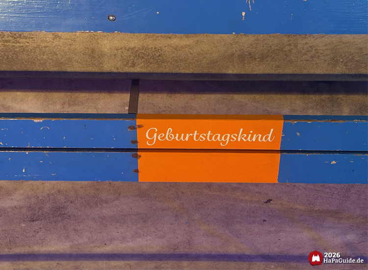 Blau gestrichene Sitzbank der Aquarena mit orange markiertem Platz und der Aufschrift Geburtstagskind