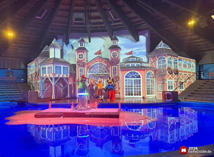 Bühne in der Aquarena des Hansa-Park mit der Familienshow Adrienne, Emmi, Pingi und Co. und die versunkene Stadt mit Wasserbecken, beleuchteter Kulisse Hansa Hotel und mehreren kostümierten Maskottchen