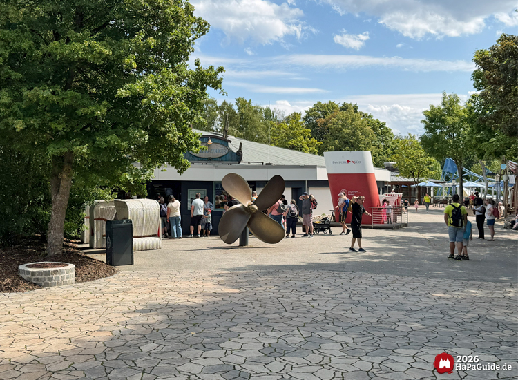 Eingangsbereich des Hansa-Park Wasser-Zirkus Aquarena mit großem Schiffspropeller, Strandkörben und Besuchenden unter blauem Himmel