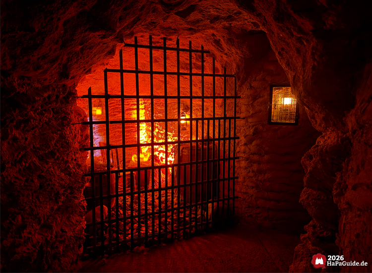 Gittertor im Inneren der Bärenhöhle im Hansa-Park mit orangefarbenem Licht und steinernen Wänden, dahinter Fässer und Laternen, vermittelt eine mystische Stimmung