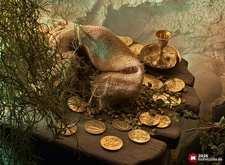 Nahaufnahme eines Schatzes in der Bärenhöhle des Hansa-Parks mit goldenen Münzen, Sackleinen und einem goldenen Kelch auf einem Holztisch, umgeben von Pflanzen