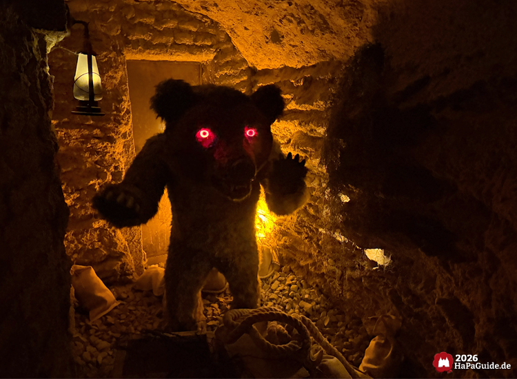 Bärenfigur mit leuchtend roten Augen in der Bärenhöhle des Hansa-Parks, steht in einer Felskammer mit Laternenlicht und Säcken auf dem Boden