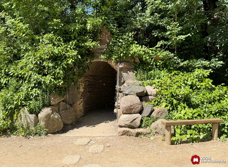 Ausgang der Bärenhöhle im Hansa-Park mit steinernem Torbogen, umgeben von dichtem Grün und großen Felsen, davor ein Weg mit Bärenspuren im Sand