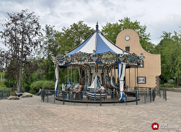 Maritimes Carrousel Baltique im Hansa-Park mit blau-weißer Dachkonstruktion, Busen und benachbarten Bronzeseelöwen, umgeben von Bäumen und dem gotischen Operator-Gebäude mit geschwungener Fassade