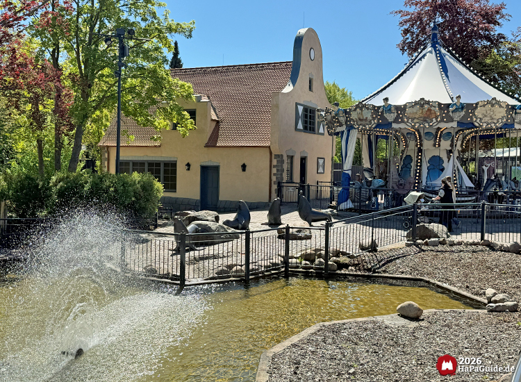 Maritimes Carrousel Baltique im Hansa-Park neben einem Teich mit Wasserfontäne und Seelöwenfiguren, dahinter das beige Karussell-Gebäude unter sonnigem Himmel
