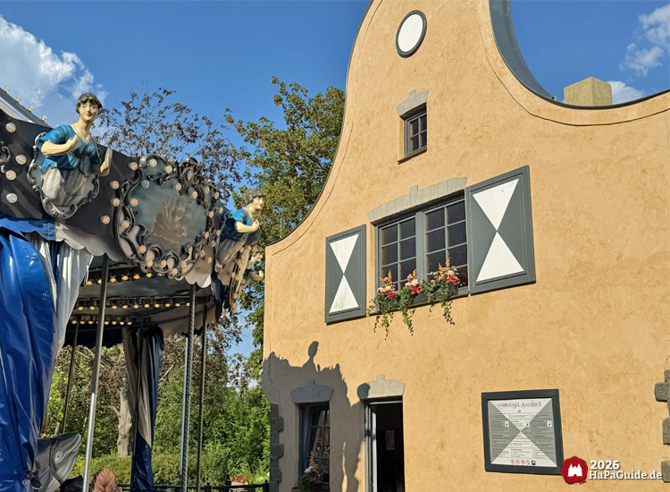 Detailansicht des maritimen Karusells mit Musen in blauen Gewändern neben einem gelb-beigen Gebäude mit grauen Fensterläden, Blumenkästen und Schild mit der Aufschrift 'Carrousel Baltique' im Hansa-Park