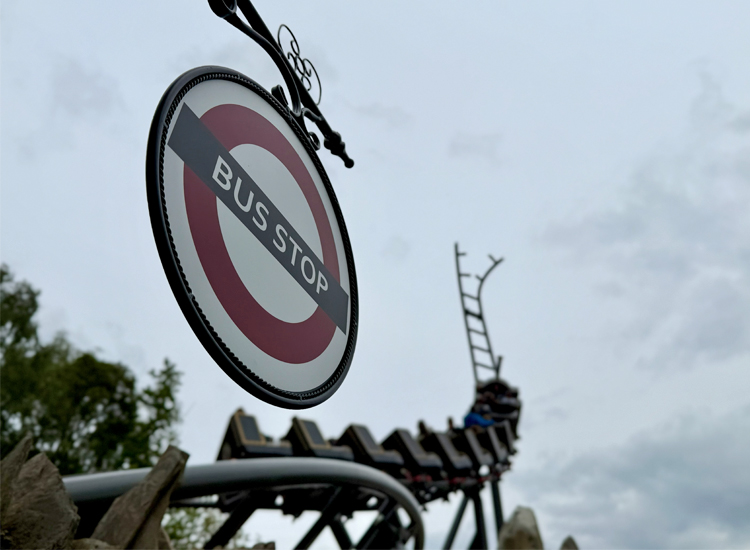 Rundes 'Bus Stop'-Schild im Hansa-Park vor der Achterbahnstrecke des Cornwall Coaster, deren Schienen in den Himmel führen, umgeben von Bäumen und grauen Wolken