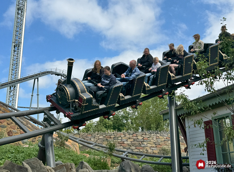 Zug der Familienachterbahn Cornwall Coaster im Hansa-Park mit schwarzer Lokomotive und goldenen Verzierungen, fährt über die Schienen mit Fahrgästen unter blauem Himmel
