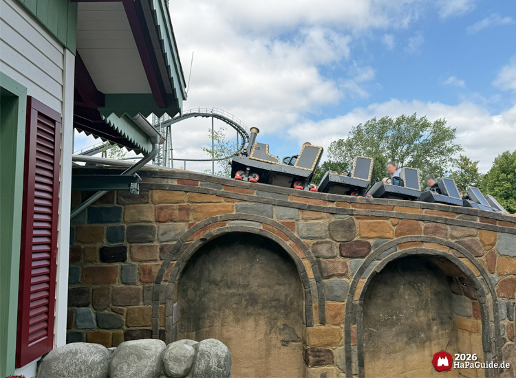 Zug der Cornwall Coaster Achterbahn im Hansa-Park fährt über ein Steinviadukt mit Rundbögen, neben einem Gebäude mit grüner Holzverkleidung und roten Fensterläden