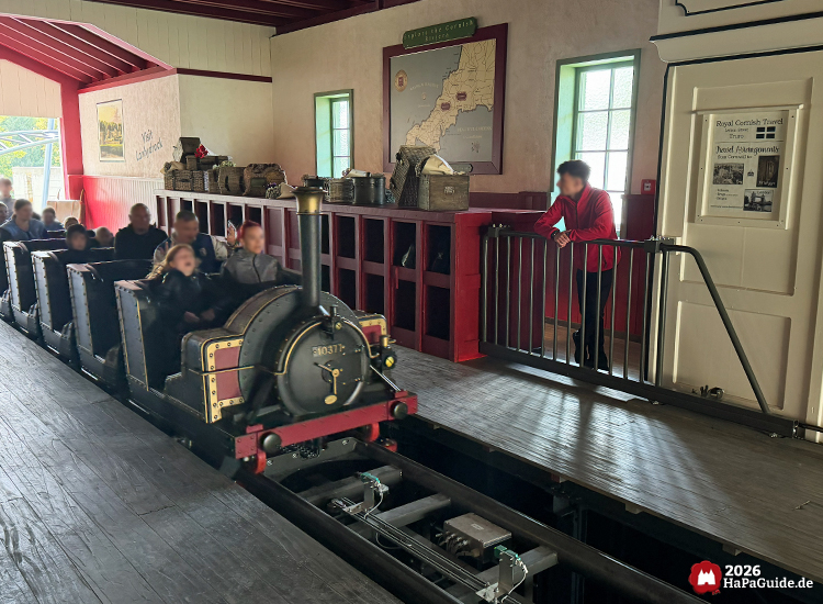 Innenbereich der Station des Cornwall Coaster im Hansa-Park mit schwarzer Lokomotive und Fahrgästen, roten Holzverkleidungen und Landkarte von Cornwall an der Wand