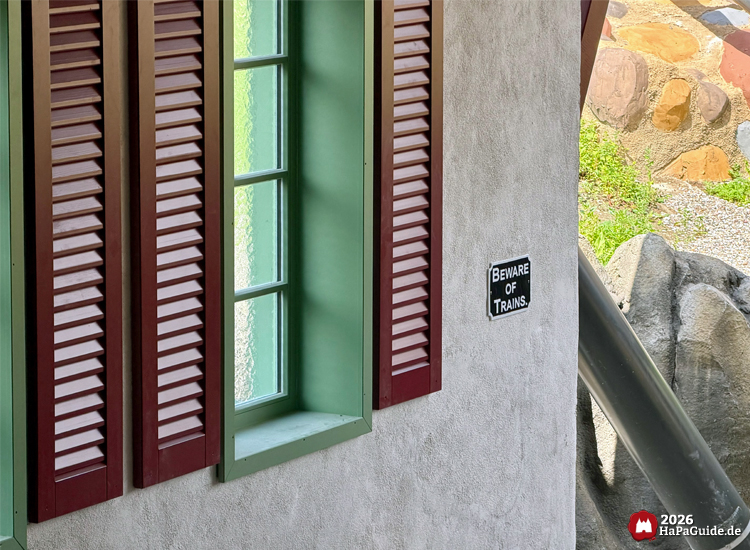 Grün gerahmtes Fenster mit roten Holzläden an der Station des Cornwall Coaster im Hansa-Park, daneben ein Schild mit der Aufschrift 'Beware of Trains'