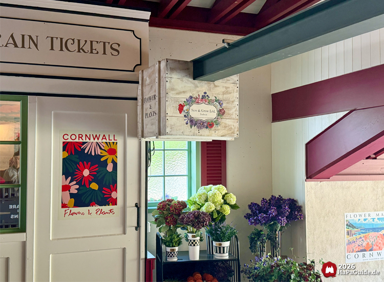 Blumendekoration im Innenbereich der Cornwall Coaster Station im Hansa-Park mit bunten Pflanzen, Holzboxen und Postern mit der Aufschrift 'Cornwall Flowers & Plants'