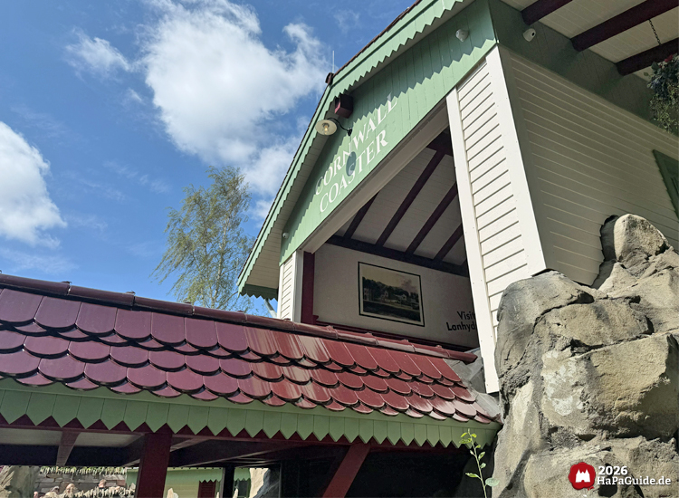 Außenansicht der Station der Cornwall Coaster Familienachterbahn im Hansa-Park mit grün-weißem Giebel, rotem Dach und Felsen, unter blauem Himmel mit weißen Wolken
