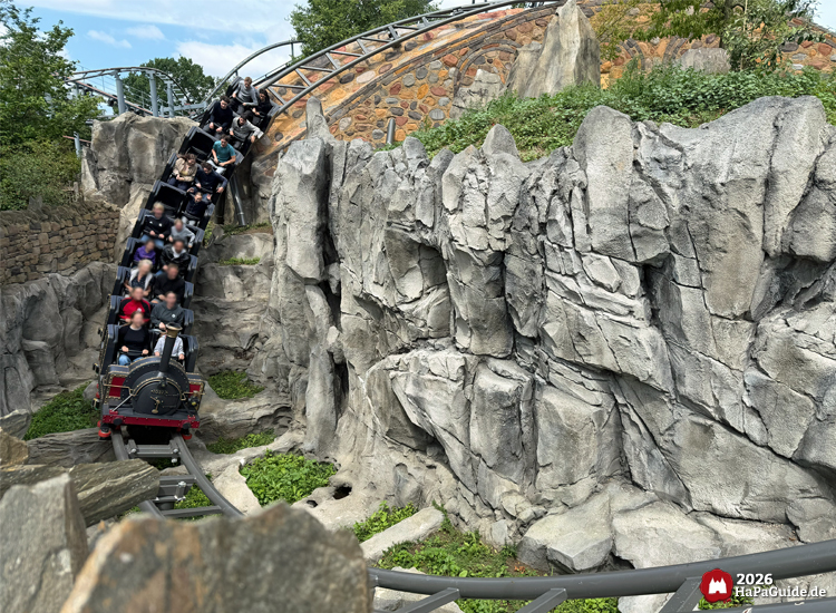 Zug der Cornwall Coaster Achterbahn im Hansa-Park fährt durch eine steile Felslandschaft mit grauen Steinwänden und grüner Vegetation, Fahrgäste genießen die Abfahrt