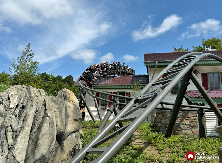 Zug der Cornwall Coaster Achterbahn im Hansa-Park fährt in einer Kurve über das Stationsgebäude mit rotem Dach und weißen Wänden, umgeben von Felsen und Pflanzen unter blauem Himmel