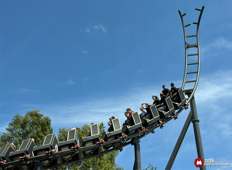 Zug der Cornwall Coaster Achterbahn im Hansa-Park fährt rückwärts auf den Spike unter blauem Himmel, Fahrgäste heben die Arme während der Fahrt, im Hintergrund grüne Bäume
