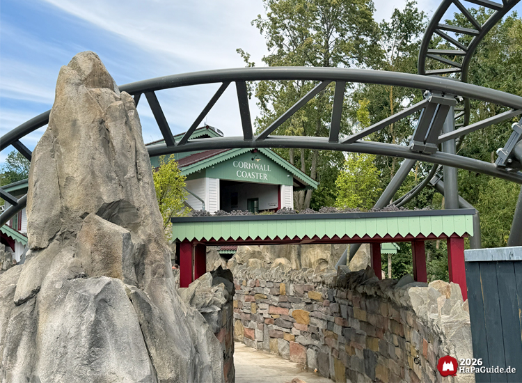 Blick vom Eingang des Wartebereichs auf die Station des Cornwall Coaster im Hansa-Park mit grün-weißem Gebäude und rotem Dach, umgeben von Felsen und der grauen Achterbahnstrecke