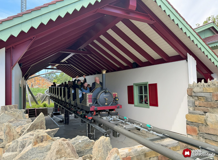 Zug der Cornwall Coaster Achterbahn im Hansa-Park fährt in den überdachten Beschleunigungsabschnitt mit weißer Fassade, grünem Fensterrahmen und roten Fensterläden, umgeben von Steinmauern