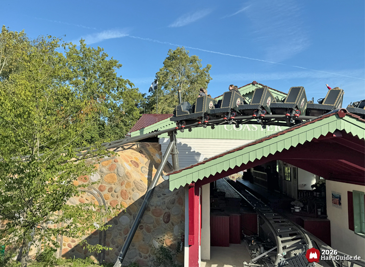 Zug der Cornwall Coaster Achterbahn im Hansa-Park fährt über das Dach der Station mit grünem Giebel und rotem Dach, dahinter Bäume und blauer Himmel mit Kondensstreifen