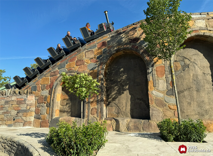 Zug der Cornwall Coaster Achterbahn im Hansa-Park fährt über eine Steinbrücke mit großen Rundbögen, umgeben von Bäumen und Pflanzen unter klarem blauem Himmel
