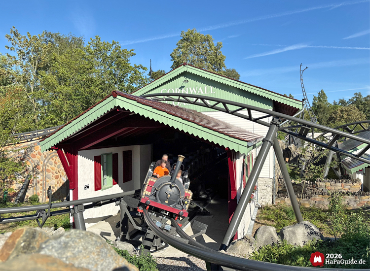 Zug der Cornwall Coaster Achterbahn im Hansa-Park verlässt die Station mit weißer Fassade und grünem Dach, schwarze Schienen führen durch Felsen und Pflanzen unter blauem Himmel