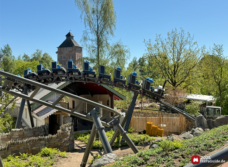 Cornwall Coaster im Hansa-Park: Achterbahn mit blauen Testdummies auf Sitzen fährt über geschwungene Schiene vor Beschleunigungsgebäude und historischem Saviour-Turm der Flucht von Novgorod, umgeben von grüner Landschaft, Foto Mai 2026 von HaPaGuide.de