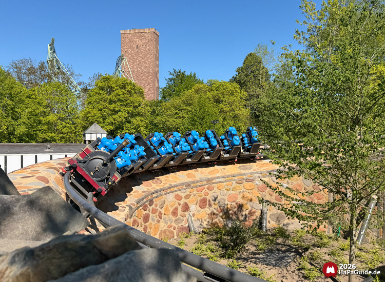 Cornwall Coaster im Hansa-Park: Achterbahnzug im Dampflokdesign mit blauen Testdummies fährt durch Kurve auf gemauerter Steinwand, umgeben von grünen Bäumen und blauem Himmel, Foto Mai 2026 von HaPaGuide.de