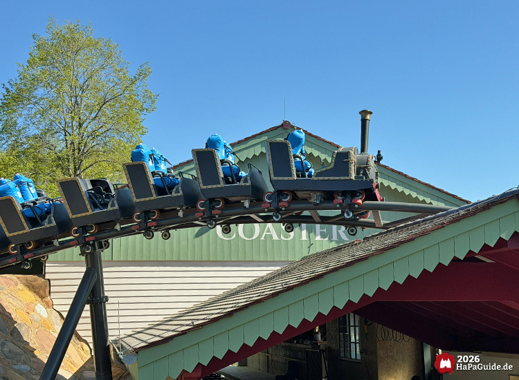 Cornwall Coaster im Hansa-Park: Achterbahnzug im Dampflokdesign mit blauen Testdummies auf Sitzen fährt über geschwungene Schiene direkt über dem Stationsgebäude mit grünem Giebel und rotem Dach, umgeben von Bäumen unter blauem Himmel, Foto Mai 2026 von HaPaGuide.de