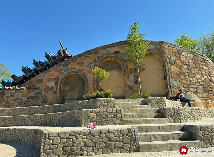 Cornwall Coaster im Hansa-Park: Achterbahnzug im Dampflokdesign mit blauen Testdummies auf Sitzen fährt über gemauertes Viadukt, davor terrassenförmige Sitzstufen und junge Bäume unter blauem Himmel, Foto Mai 2026 von HaPaGuide.de