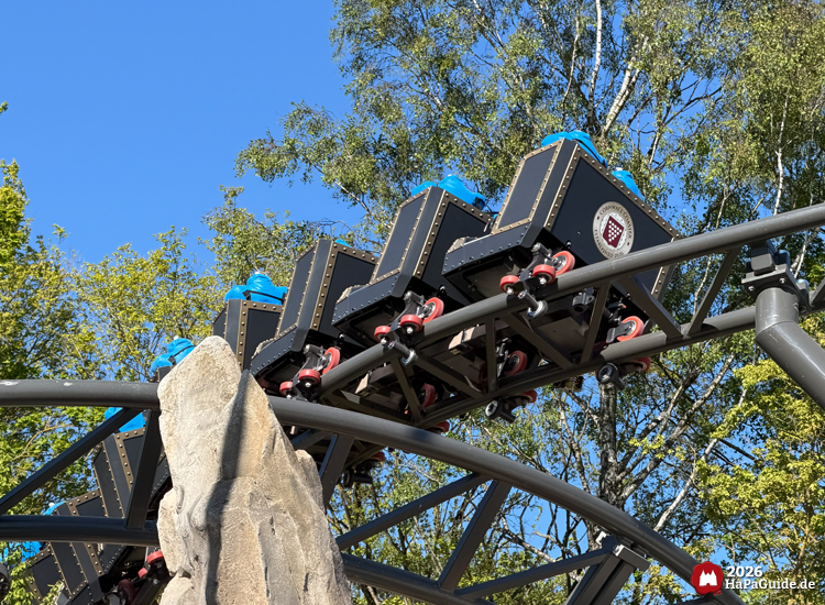 Cornwall Coaster im Hansa-Park: Achterbahnwagen im schwarzen Design mit blauen Testdummies fährt über geschwungene Schiene zwischen Felsen und grünen Bäumen unter klarem Himmel, Foto Mai 2026 von HaPaGuide.de