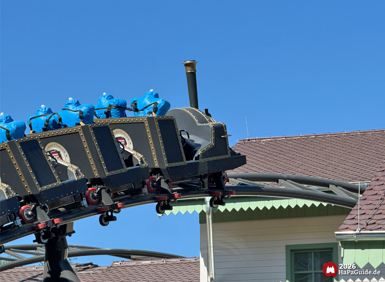 Cornwall Coaster im Hansa-Park: Achterbahnzug im Dampflokdesign mit blauen Testdummies fährt über geschwungene Schiene direkt am hellen Stationsgebäude mit grünem Dachrand unter klarem Himmel, Foto Mai 2026 von HaPaGuide.de