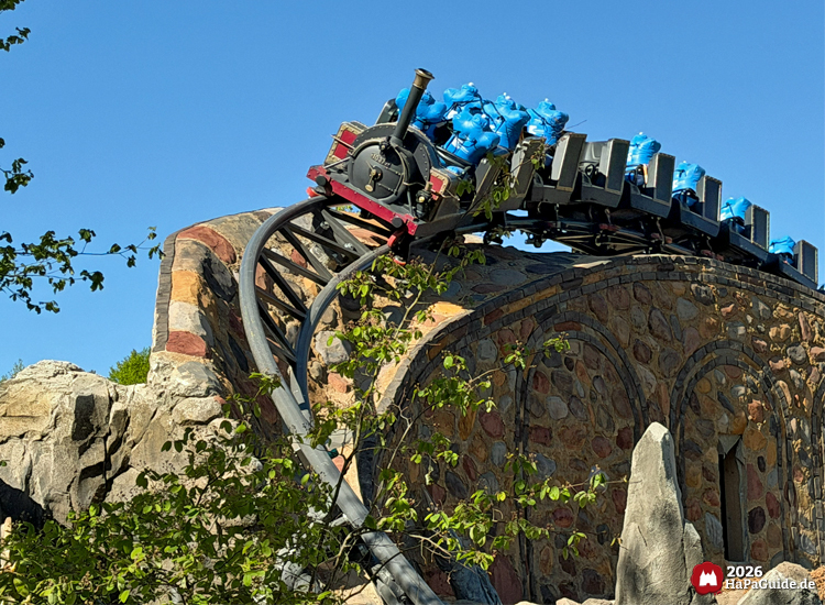 Cornwall Coaster im Hansa-Park: Achterbahnzug im Dampflokdesign mit blauen Testdummies auf Sitzen fährt über geschwungene Schiene auf gemauertem Viadukt, umgeben von Felsen und grüner Vegetation unter blauem Himmel, Foto Mai 2026 von HaPaGuide.de