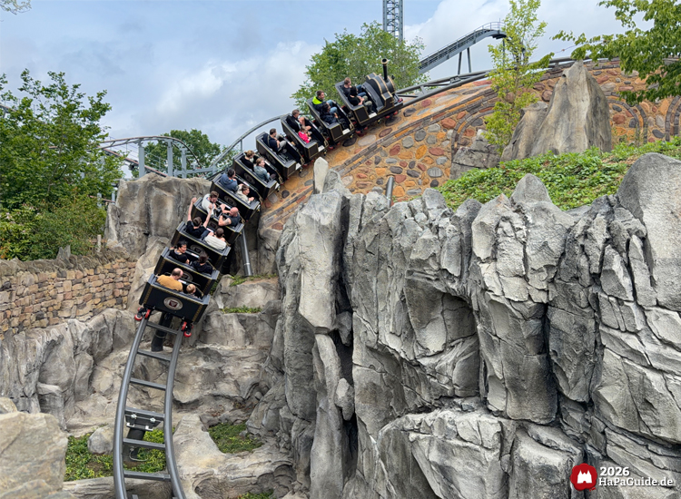 Cornwall Coaster im Hansa-Park mit schwarzen Wagons fährt eine steinerne Brücke hinunter durch felsige Landschaft, umgeben von grünen Bäumen unter wolkigem Himmel