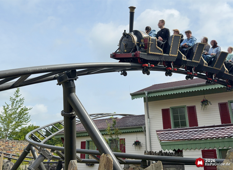 Schwarze Dampflokomotive als Achterbahnzug des Cornwall Coaster im Hansa-Park fährt über eine geschwungene Stahlstrecke vor einem hellen Gebäude mit roten Fensterläden und grünem Dachrand unter blauem Himmel