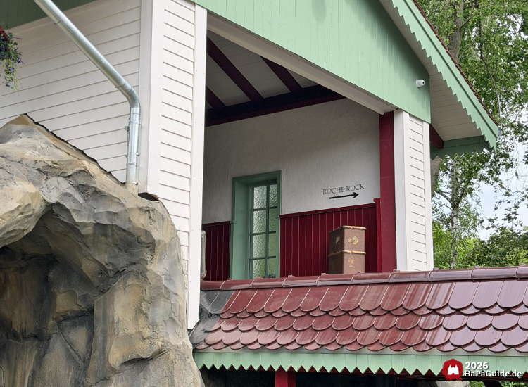 Detailansicht des hellen Bahnhofsgebäudes vom Cornwall Coaster im Hansa-Park mit grünem Dachrand und roten Balken, daneben eine Felsformation; im Inneren ein grünes Fenster, eine braune Truhe und ein Schild mit der Aufschrift 'Roche Rock' mit Pfeil nach rechts