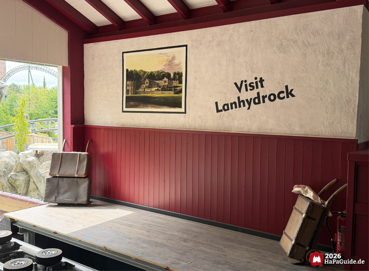 Thematisierte Bahnsteigszene im Bahnhof des Hansa-Park Cornwall Coaster mit roter Holzverkleidung, Koffern und einem Wandbild von Lanhydrock; daneben der Schriftzug 'Visit Lanhydrock' und im Hintergrund Felsen, Pflanzen und Teile der Achterbahnstrecke