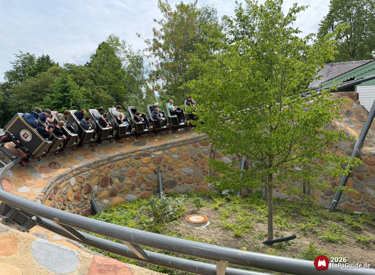 Cornwall Coaster im Hansa-Park mit schwarzen Wagons fährt eine geschwungene Steinmauer entlang, umgeben von grünen Bäumen und Sträuchern unter leicht bewölktem Himmel