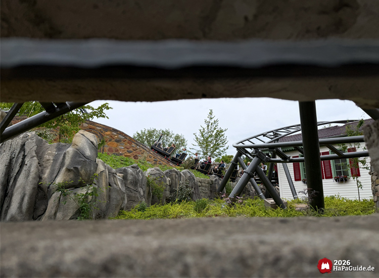 Blick durch eine Felsöffnung auf die Achterbahn Cornwall Coaster im Hansa-Park, die zwischen Felsen und grüner Vegetation entlang eines weißen Gebäudes mit roten Fensterläden fährt