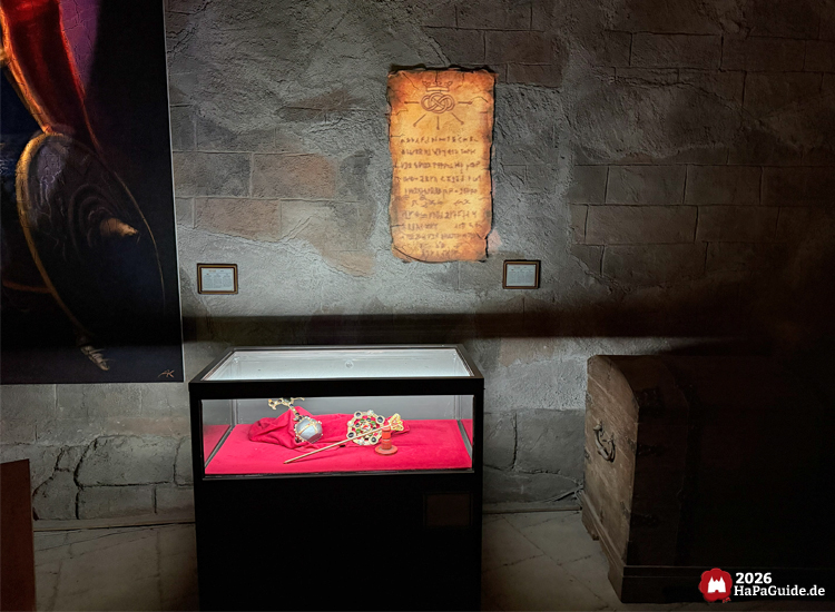 Ausstellungsbereich im Kärnan Museum im Hansa-Park mit beleuchteter Vitrine, in der goldene und verzierte Artefakte auf rotem Stoff präsentiert werden, daneben eine alte Schriftrolle an der Steinwand