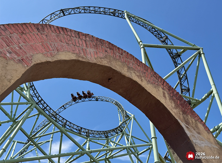 Achterbahn Der Schwur des Kärnan im Hansa-Park mit grünem Herzelement, fotografiert durch Backsteinbogen unter blauem Himmel, dynamische Perspektive