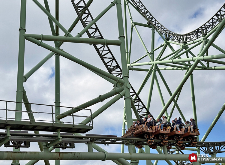 Zug der Achterbahn Der Schwur des Kärnan im Hansa-Park fährt entlang grünem Stahlgerüst, Fahrgäste in braunen Sitzen unter blauem Himmel mit Wolken