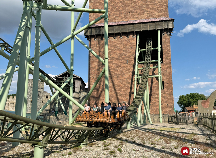 Achterbahn Der Schwur des Kärnan im Hansa-Park mit Zug vor dem hohen Kärnan-Turm aus Backstein, grüne Stahlträger und mittelalterliche Kulisse im Hintergrund