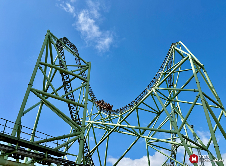 Achterbahn Der Schwur des Kärnan im Hansa-Park mit Zug auf höchstem Punkt der Strecke, grüne Stahlkonstruktion vor blauem Himmel mit wenigen Wolken