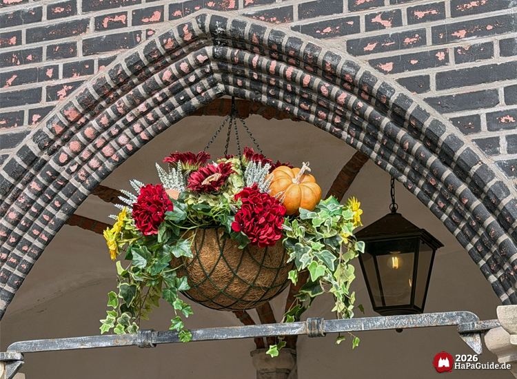 Herbstliche Blumenampel mit Kürbis, roten und gelben Blüten unter gemauertem Spitzbogen in der Hanse in Europa im Hansa-Park, dahinter schwarze Laterne