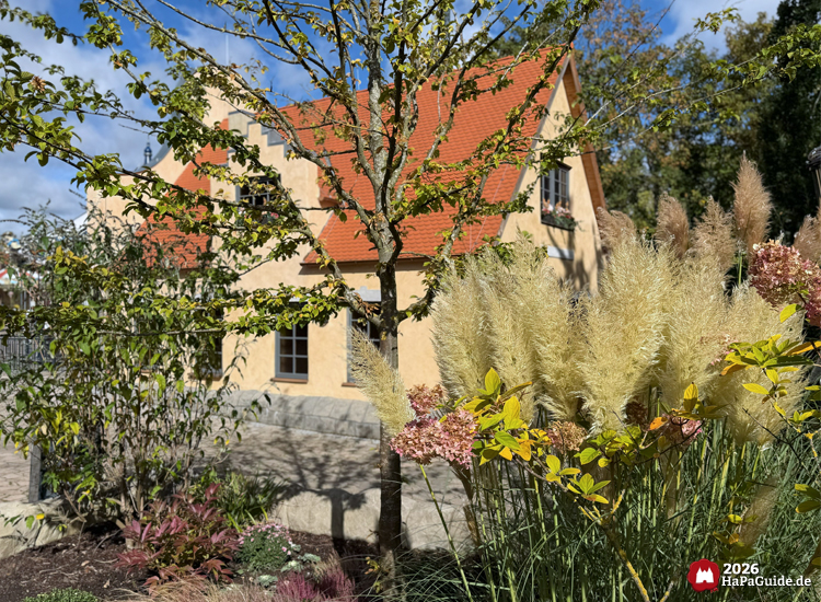 Gelbes Haus mit rotem Dach im Hansa-Garten des Hansa-Park, umgeben von Garten mit Pampasgras, Hortensien und herbstlicher Bepflanzung unter blauem Himmel mit weißen Wolken