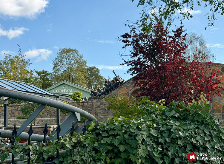 Achterbahn Cornwall Coaster im Themenbereich Bezauberndes Britannien des Hansa-Parks mit Zug im Stil einer alten Dampflokomotive, umgeben von grüner und roter Vegetation, sonniger Herbsttag mit blauem Himmel