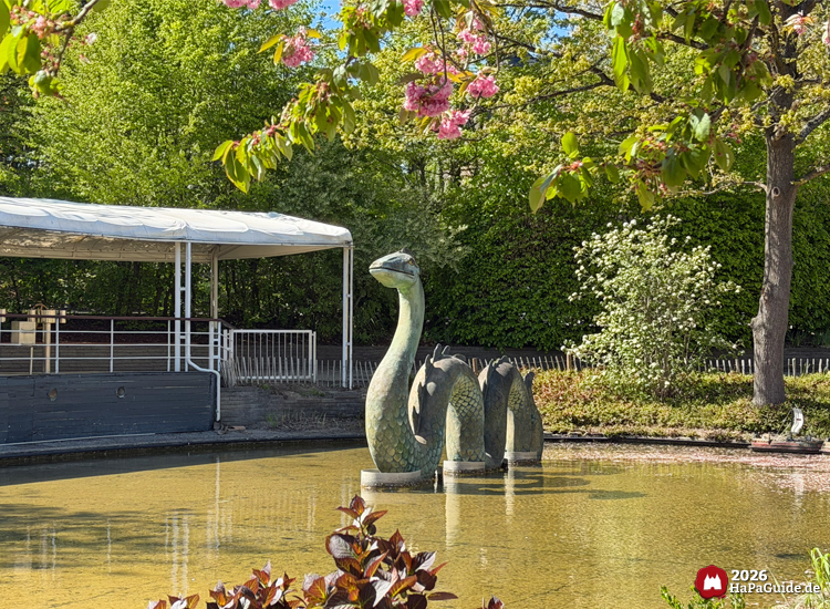 Grüne Seeschlange Nessie im Teich der Fernlenkschiffe des Hansa-Parks unter blühenden Kirschzweigen, umgeben von frischem Frühlingsgrün und einem überdachten Steg im Hintergrund